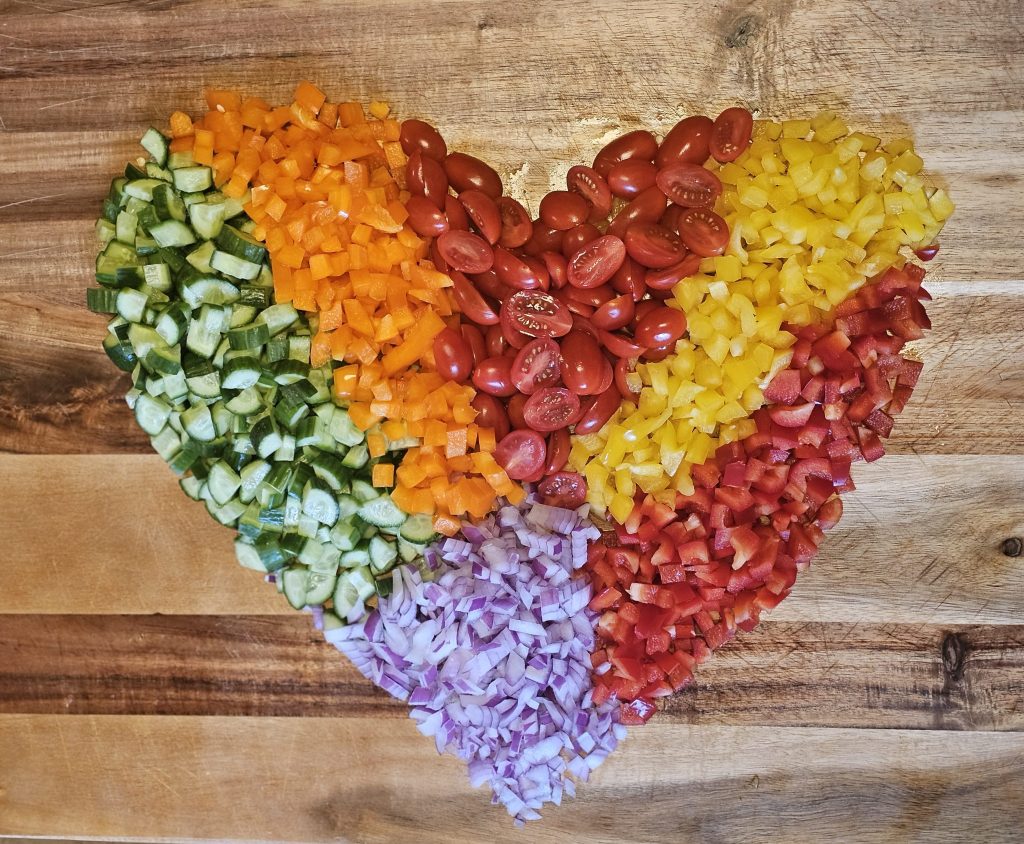 A heart-shaped arrangement of colorful chopped vegetables on a wooden surface, including green cucumbers, orange bell peppers, red tomatoes, yellow bell peppers, and purple onions.