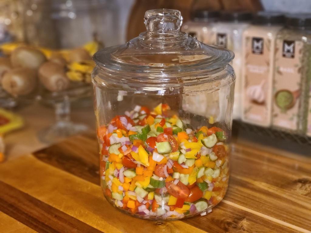 A glass jar filled with colorful chopped vegetables including bell peppers, cucumbers, red onions, and cherry tomatoes, placed on a wooden countertop.